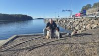 En man sitter vid vattenkanten tillsammans med två hundar på en grusig strand.