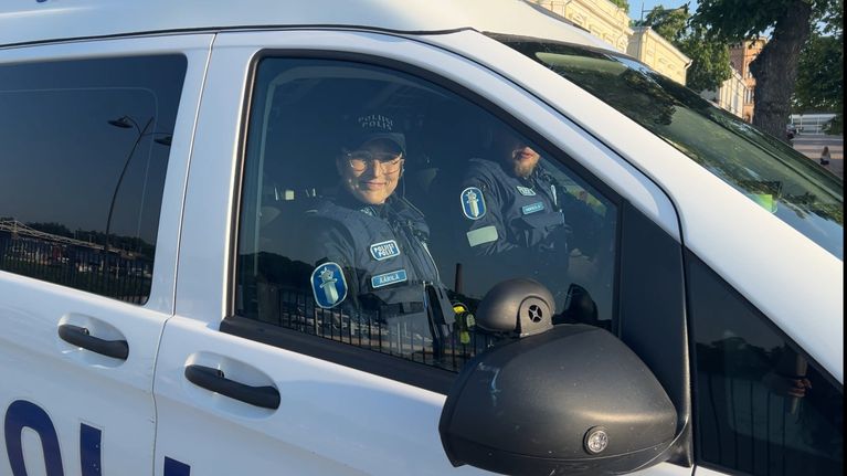 Hymyilevä naispoliisi, vanhempi konstaapeli Mervi Äärilä, poliisauton etupenkillä, kuskina parrakas nuori mies.