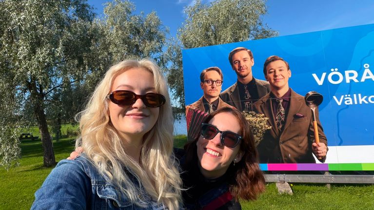 Två personer står framför en skylt med tre män och texten "VÖRÅ Välko". Träd och gräs syns i bakgrunden.