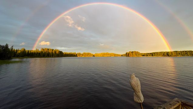 Järven yllä näkyy kirkas sateenkaari ja sen yläpuolella himmeämpi, etualalla puiselta laiturilta erottuu lintupatsas.