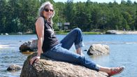 En kvinna med långt, grått hår sitter på en stor sten vid havet. Hon är klädd i svart ärmlös tröja, blå jeans och röda sneakers. I bakgrunden hav och skog. 