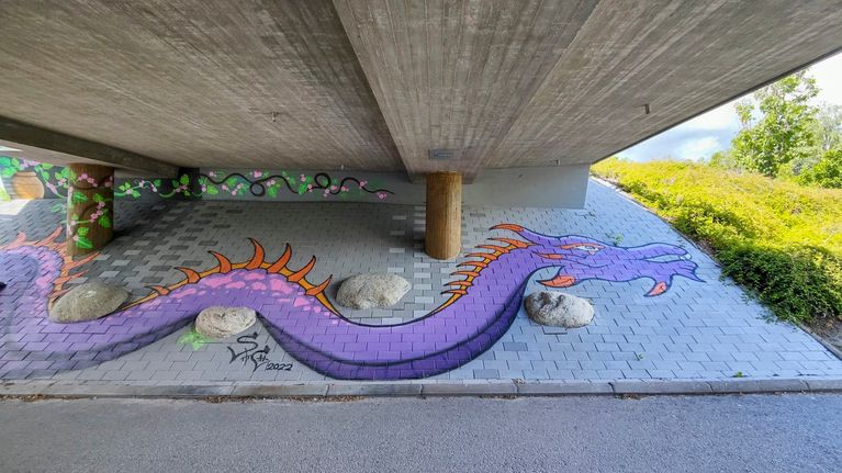 Alikulkutunnelin harmaaseen seinään maalattu violetti lohikäärme-graffiti.
