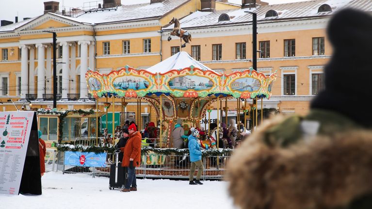 Karuselli on lumessa Tuomaan markkinoilla ja ihmisiä seisoo karusellin edessä. Etualalla ihminen hupussa.