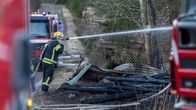 Pelastaja sammuttaa maastopaloa vedellä. Maassa on esimerkiksi peltiä. Kuvassa näkyy myös pelastusajoneuvoja.