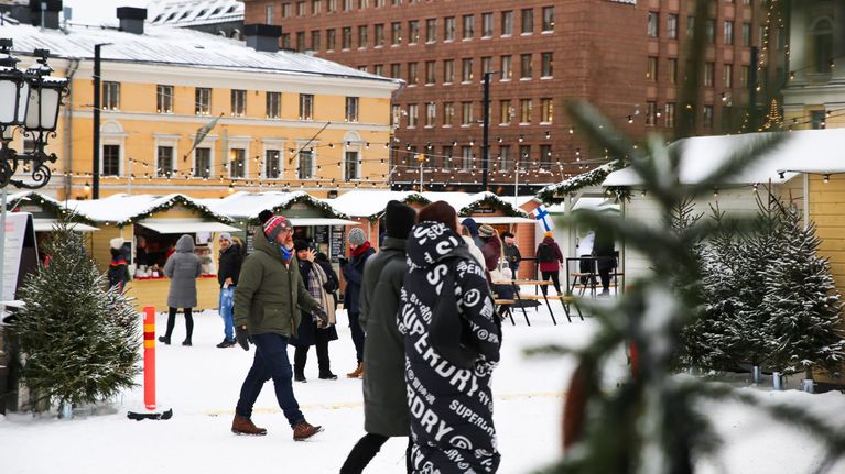 Ihmiset kävelevät Tuomaan markkinoilla kuusien ja kojujen keskellä. Maassa on lunta.