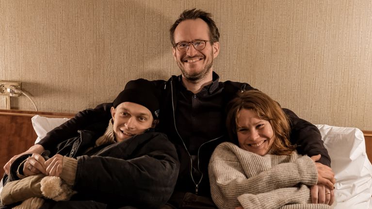Juho Kuosmanen sitter i en säng och håller om huvudrollsinnehavarna, skådespelarna Juri Borisov och Seidi Haarla.