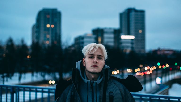 Vaaleahiuksinen mies seisoo sillalla iltavalossa, taustalla näkyy kerrostalojen siluetteja ja autojen valoja.