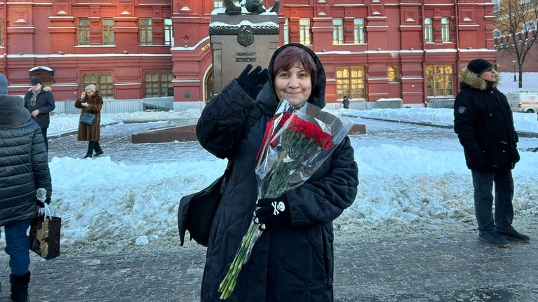 Darja Mitina vilkuttaa oikealla kädellään pidellen toisessa kädessään muoviin käärittyjä ruusuja. Taustalla näkyy marsalkka Georgi Zhukovin ratsastajapatsas.