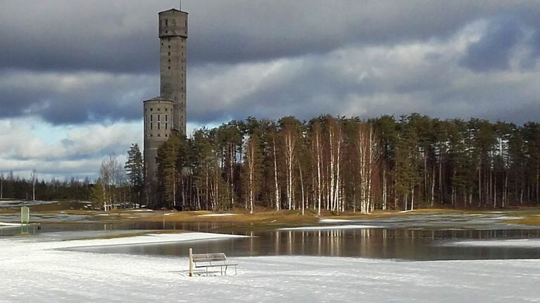 Edessä penkki veden äärellä. Takana kohoaa Keretin kaivostorni Outokummussa. 