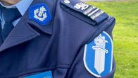 Poliisin univormu, jossa näkyy arvomerkkejä, hihan vaakuna ja osa rinnassa olevaa nimikylttiä, taustalla nurmikkoa.