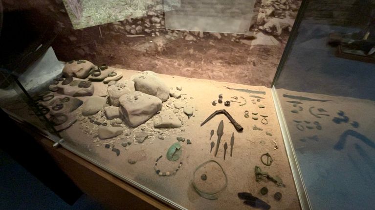 Satakunnan museon vitriini, jossa on esillä erilaisia historiallisia esineitä, kuten koruja, työkaluja, jotka on asetettu hiekkaiselle alustalle.