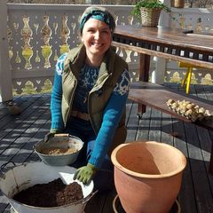 En kvinna sitter på huk på en veranda med en stor kruka, en liten zinkhink och en emaljerad balja med trädgårdsjord.