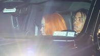 Auton etuistuimilla istuu kaksi henkilöä, toinen katsoo eteenpäin ja toisella on oranssi tukka. Auton kojetaululla on tarroja ja papereita.