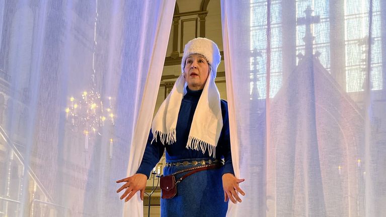 Valkeaan hattuun ja siniseen asuun pukeutunut nainen avaa vaaleat verhot kirkon alttarilla.
