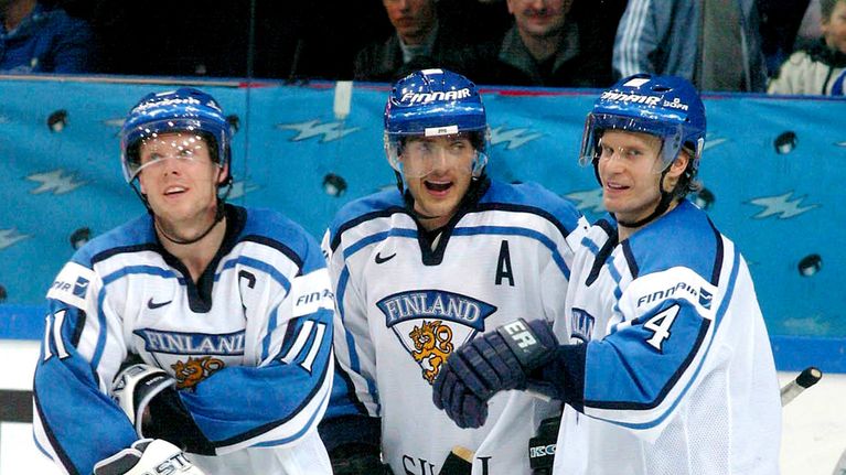 Saku Koivu, Teemu Selänne ja Kimmo Timonen syleilevät valkoisissa maajoukkuepaidoissa jääkiekkokentällä.