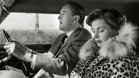 Mies (George Sanders) ajaa autoa, matkustajan paikalla istuu nainen (Ingrid Bergman) leoparditurkissa ja selaa matkaopasta. Kuva elokuvasta Matka Italiaan (Matka Italiassa, Viaggio in Italia).