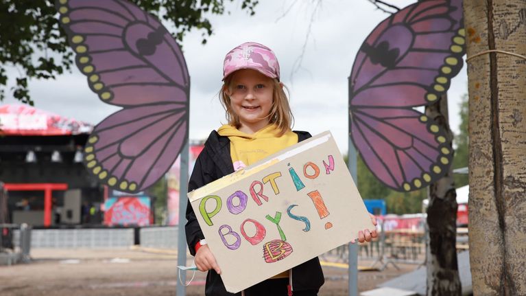 Seitsemänvuotias Eini Nalli pitää käsissään kylttiä, jossa teksti Portion Boys, taustalla violetit perhosen siivet.