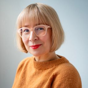 Tiina-Rakel Liekki katsoo kameraan. Hänellä on polkkatukka, silmälasit ja oranssi villapaita.