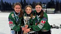 Tre idrottare i gröna tröjor firar med en pokal på isen under kvällstid.