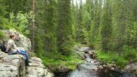 En ung man sitter vid en brant klippa i skogen invid en liten älv.