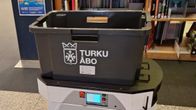 En robot med en svart låda märkt ”Turku Åbo” står framför bokhyllor i ett bibliotek.
