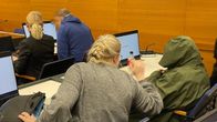 Flera personer sitter i en rättssal. En gömmer sig bakom under en grön jacka och framför henne sitter en man i blå munkjacka med pixelerat ansikte.