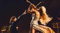 A man in black stands with outstretched arm as a woman with long blonde hair plays a violin above him, seen from behind.