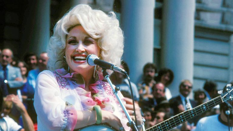 Dolly Parton ler och tittar åt sidan. Hon håller i en gitarr vid en mikrofon.