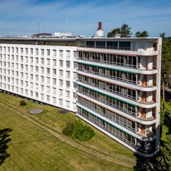 Vaalea usean kerroksen rakennus metsän ja nurmialueen reunalla, parvekkeita ja ikkunoita julkisivussa.