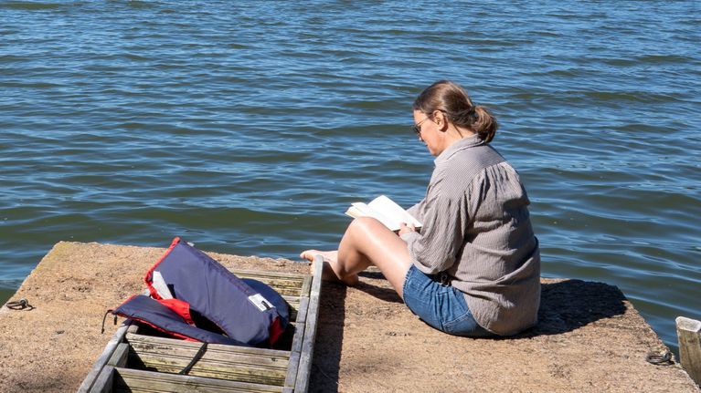 En person sitter på en brygga vid en sjö och läser en bok; en ryggsäck ligger bredvid.