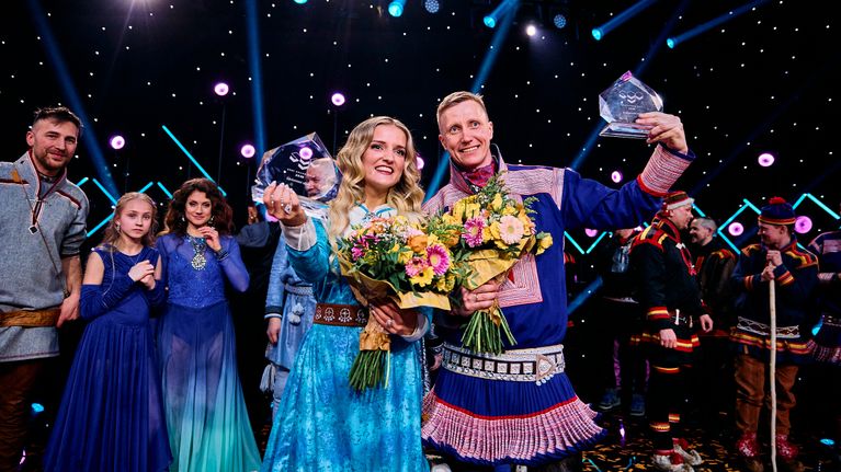 Ellinor Halvari ja Ánte Niillas Bongo lavalla Sámi Grand Prix palkintojen kanssa. Taustalla näkyvät muut kilpailuun osallistuneet.