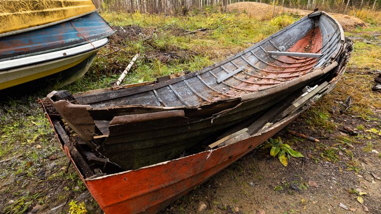 Kuvassa näkyy vanha, huonokuntoinen puuvene, joka on jätetty maalle lojumaan. Veneessä on ohut, rikkoutunut lasikuitupinnoite.