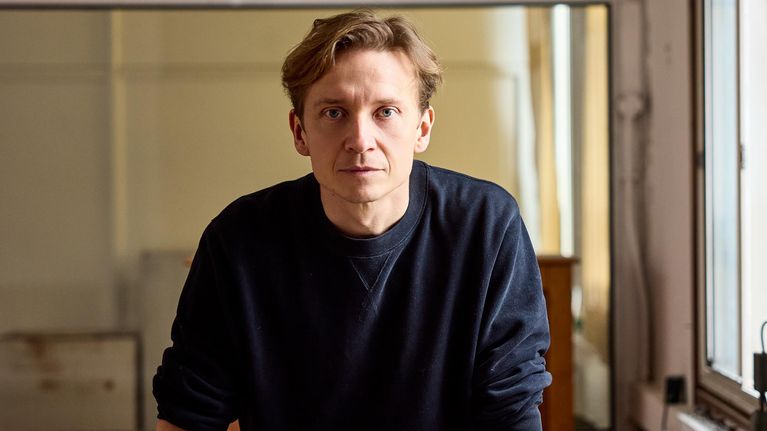 En blond man står vid ett bord med olika skulpturdelar och modeller i ett ljust rum.
