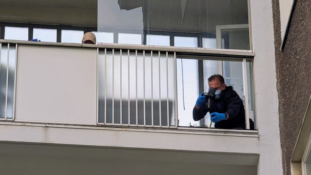A man wearing dark clothing, blue gloves and a face mask photographing a broken balcony, seen from below.