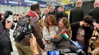 Two women embracing at an airport, one holding a small Palestinian flag, with other people around them.