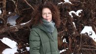 En person med mörkt lockigt hår och grön jacka står framför en rotvälta med inslag av snö.