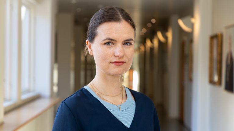 En person med mörkt hår och blå topp står i en ljus korridor med fönster och tavlor på väggarna.