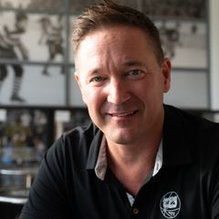 Mies istuu pöydän ääressä ravintolassa, taustalla jääkiekkoaiheinen valokuvaseinä.
