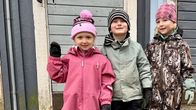 Tre barn i overaller, mössor och vantar står utomhus framför en blågrå vägg.