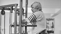 A black-and-white photograph shows a stocky, moustached man standing at a gym rack.