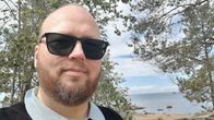 En person med skägg och solglasögon står utomhus vid en strand med träd, stenar och hav i bakgrunden.
