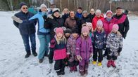 En grupp människor står tillsammans utomhus på en snötäckt mark, klädda i vinterkläder och tittar mot kameran. Det är en blandning av vuxna och barn, med träden och ett vintrigt landskap i bakgrunden.