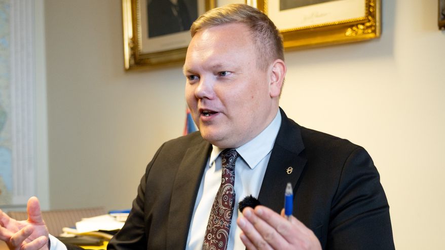 Pukuun pukeutunut henkilö keskustelee toimistossa, taustalla näkyy muotokuvia seinällä.