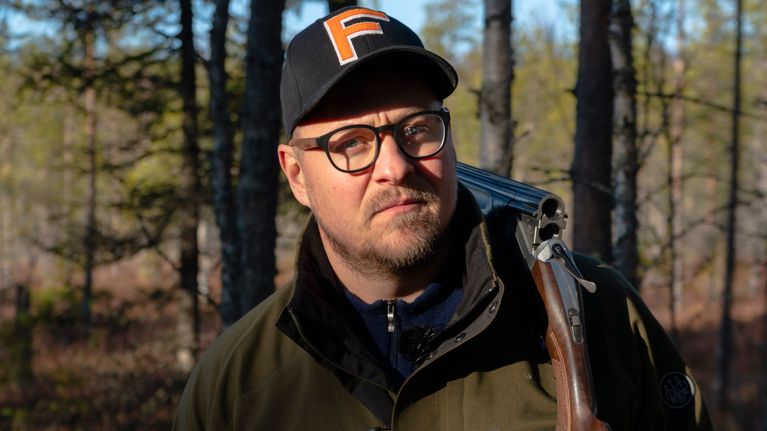 Mies seisoo metsässä teeri kädessä ja haulikko olalla.