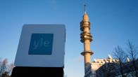 Yles logo i förgrunden på en skylt, i bakgrunden Yletornet. Solen skiner.