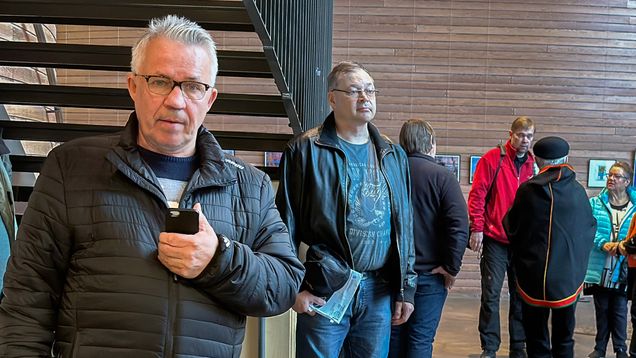 Mies seisoo portaiden alapuolella puhelin kädessään. Taustalla Saamelaiskulttuurikeskus Sajoksen aulassa seisoo muita ihmisiä.