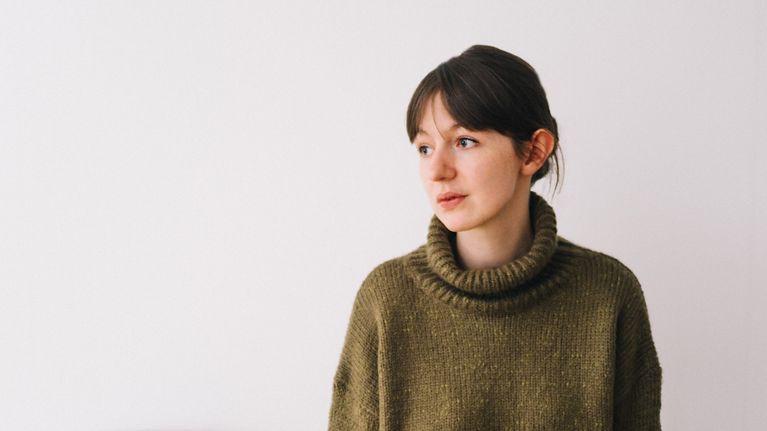 Kirjailija Sally Rooney nojaa puiseen pöytään. Hänellä on päällään villapaita ja hame.
