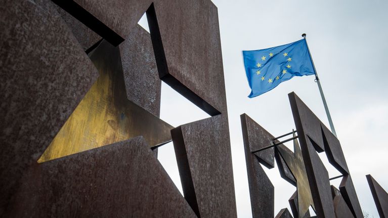 Kolmeen teräslevyyn on leikattu tähtiä ja taustalla liehuu Euroopan unionin lippu.