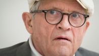 En äldre person med vit keps och glasögon syns i närbild mot en ljus bakgrund.