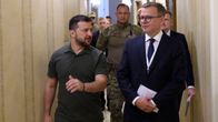 Volodymyr Zelenskyj och Petteri Orpo går bredvid varandra i en korridor. De tittar mot varandra och talar. I bakgrunden syns fler personer, bland annat en man klädd i militära kläder.
